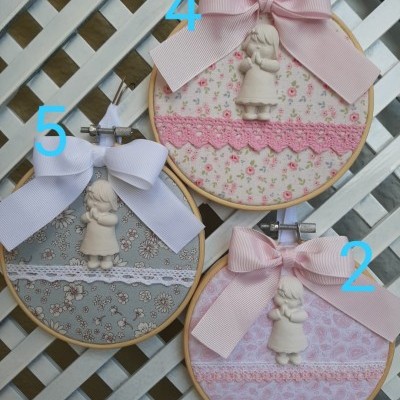 Três bastidores de bordado em madeira com tecidos floridos e laços, decorados com figuras brancas de menina