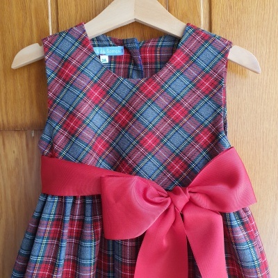 Vestido infantil xadrez vermelho e azul com laço vermelho pendurado num cabide de madeira
