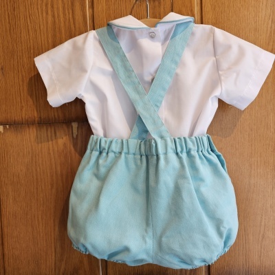 Roupa de bebé com camisa branca e calções azuis com suspensórios pendurada