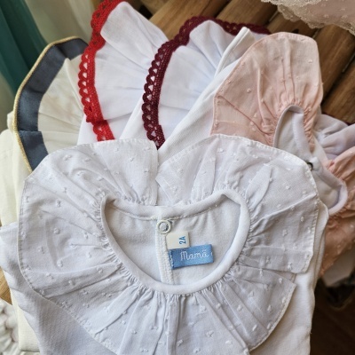 Babetes de bebé em tecidos diversos com rendas coloridas e textura de pequenos relevos, com etiquetas visíveis, em fundo de madeira.