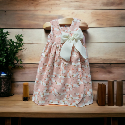 Vestido infantil rosa com padrão floral e laço branco pendurado num cabide de madeira.