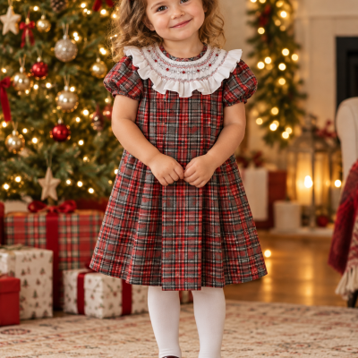 Menina com vestido xadrez vermelho e cinza, gola branca, collants brancos e sapatos castanhos, junto a árvore de Natal decorada.