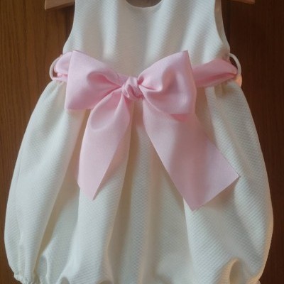 Vestido de bebé branco com cinta rosa e laço grande pendurado numa porta de madeira