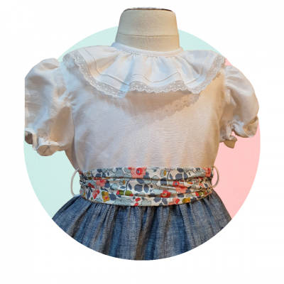 Blusa branca com gola de renda, saia azul e cinto floral colorido num manequim