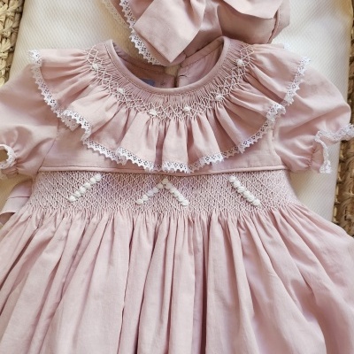 Vestido e chapéu de bebé rosa com folhos e bordados brancos numa moisés de palha.