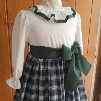 Vestido infantil com parte superior branca, gola de babado com fita verde e saia plissada xadrez azul e verde com cinto verde e laço