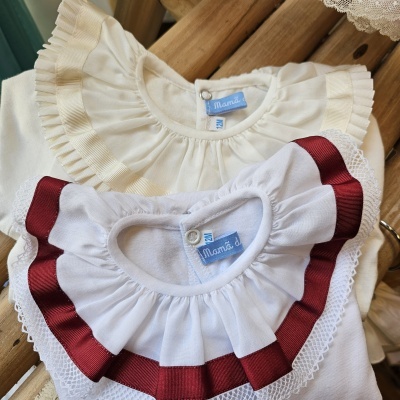 Dois bodies de bebé com gola franzida, um creme e outro branco com detalhes de fita e renda, etiquetas azuis, sobre madeira.