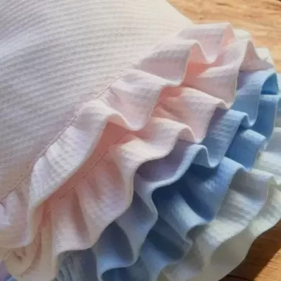 Toucas para banho em branco, rosa e azul com babados, sobre superfície de madeira.