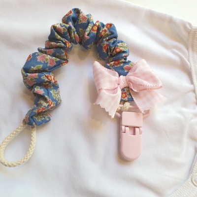 Prendedor para chupeta azul com padrão floral e laço rosa sobre tecido branco