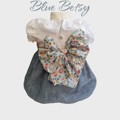 Vestido infantil branco e azul com grande laço floral e texto Blue Betsy no fundo