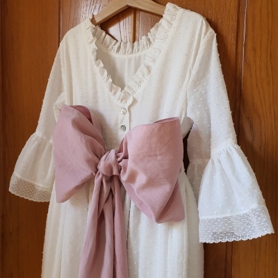 Vestido branco texturizado com laço rosa na cintura, pendurado numa porta de madeira.