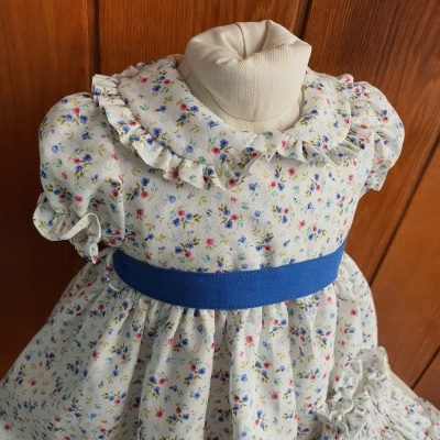 Vestido infantil branco com padrão floral e cinto azul num manequim
