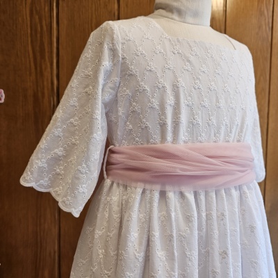 Vestido branco bordado com faixa rosa em manequim com fundo de madeira