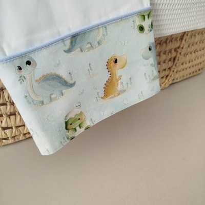 Lençol branco com bainha azul clara com estampado de dinossauros coloridos em cama com vime e tecido branco acolchoado
