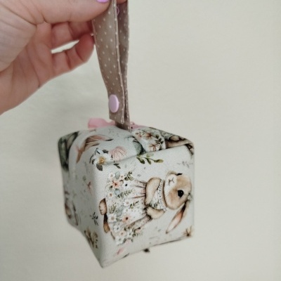 Produto de tecido em forma de cubo com padrão de coelhos e flores, alça castanha com bolinhas brancas e botão rosa