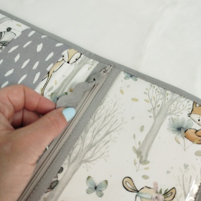 Capa de tecido plástico transparente com padrão de animais e árvore, com fecho de correr aberta por mão com unhas azuis