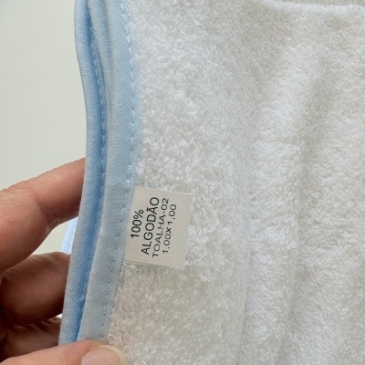 Toalha branca de algodão com borda azul clara e etiqueta visível, segurada por uma mão.