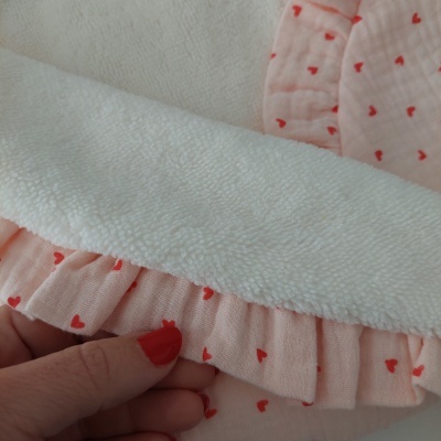 Toalha branca felpuda com tecido rosa e corações vermelhos, segurada por mão com unhas pintadas de vermelho.