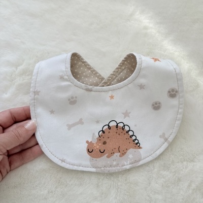 Babete de tecido branco com desenho de dinossauro castanho e estrelas, segurado por uma mão