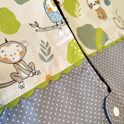 Tecido para bebé com padrão de animais e folhas, borda ondulada verde, tecido cinzento com bolinhas brancas e fecho de botão branco.