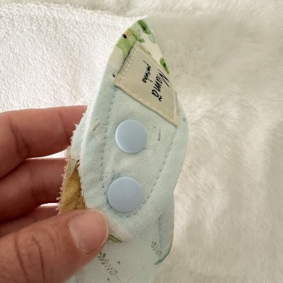 Pano de tecido azul com padrão floral, botão azul e etiqueta Katana paninho numa superfície branca felpuda.
