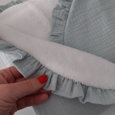 Cobertor azul claro com interior de lã branca e borda de folhos, segurado por mão com unhas vermelhas