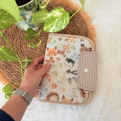Capa em tecido com desenhos de animais da floresta e pala riscada com botão, entre plantas sobre superfície branca.