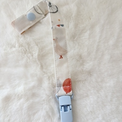 Presilha para chupeta branca com estampas coloridas e clip azul claro sobre superfície branca felpuda