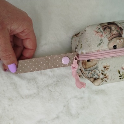Mala pequena em tecido com padrão floral e coelhos, fecho rosa e uma mão segura a alça bege com pontos brancos.
