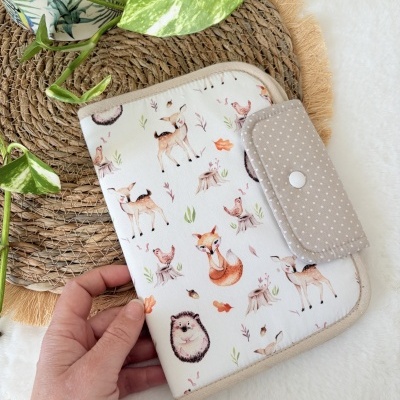 Protector de tecido com padrão de animais da floresta e fecho em botão branco sobre tapete de palha.