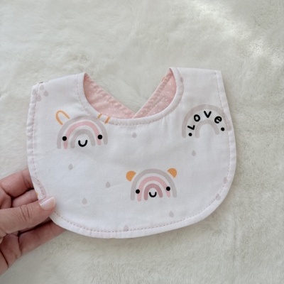 Babete para bebé branco com padrões de arco-íris e interior rosa em fundo branco felpudo