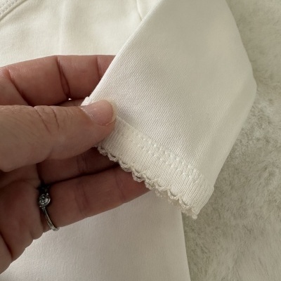 Peça de roupa branca de algodão com etiqueta 0M, bainha decorativa, segurada por uma mão.