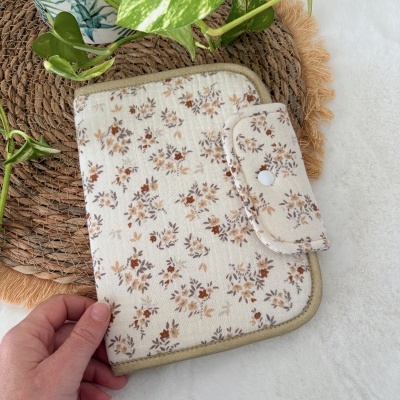 Carteira em tecido branco com padrão floral castanho e bege, botão de pressão, sobre tapete e planta.