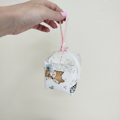 Saco pequeno de tecido branco com estampas de cães e flores, segurado por uma mão contra parede clara.