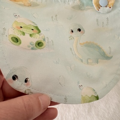 Babete infantil com desenhos de dinossauros e sapos em tons pastel.