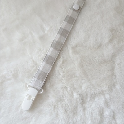 Prendedor de chupeta xadrez cinza e branco com clip branco sobre tecido felpudo branco.