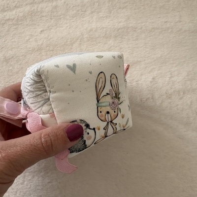 Estojo tecido branco com estampa coelho e ouriço, fecho rosa segurado por mão com unha rosa