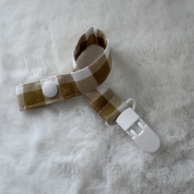 Presilha para roupa de bebé xadrez castanho e branco com mola e botão branco sobre tecido cinzento claro