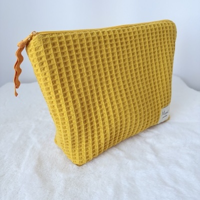 Necessaire de tecido amarelo mostarda com padrão quadriculado em relevo e fecho de correr com puxador em fita ondulada cor de laranja.