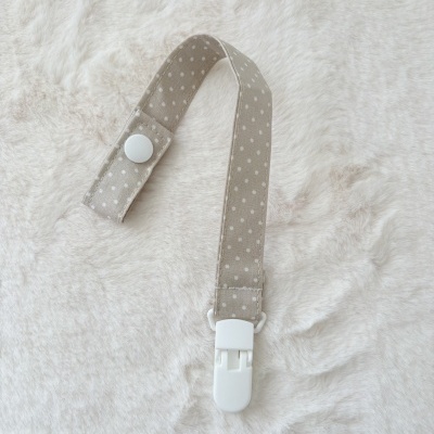 Suporte para chupeta bege com bolinhas brancas e clip branco sobre fundo felpudo branco