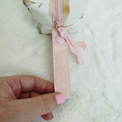 Necessaire branca com padrão castanho e rosa e fecho rosa segurada por mão com unhas cor-de-rosa