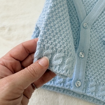 Camisola de malha azul bebé com botões e padrão tricotado a ser segurada por uma mão.