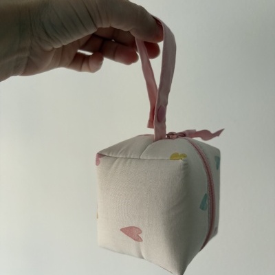 Bolsa quadrada de tecido com padrão de corações coloridos e fecho rosa sendo segurada por uma mão