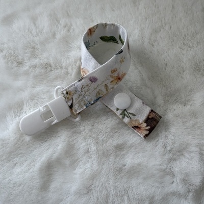 Correia para chupeta branca com padrão floral e fecho branco sobre fundo felpudo cinza claro