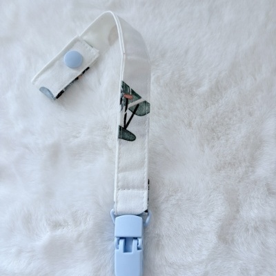 Prendedor de chupeta com fita branca estampada e clip azul claro em fundo branco felpudo