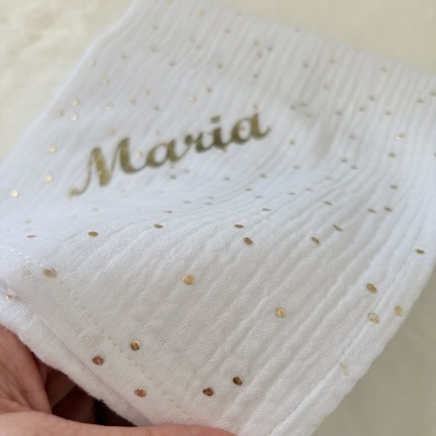 Pano branco com pontos dourados e o nome Maria impresso em dourado, segurado por uma mão.