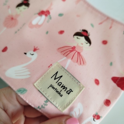 Pano rosa com estampa de bailarinas e cisnes, etiqueta Mamã pintinha e mão com unha rosa