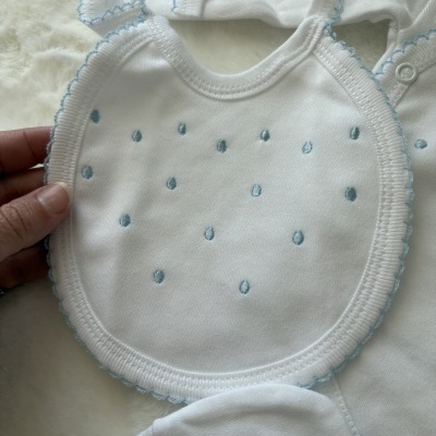 Babete branco para bebé com bordados azuis e roupa branca ao fundo.