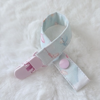 Prendedor para chupeta azul claro com botão e fecho rosa em fundo branco felpudo.