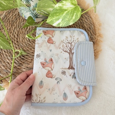 Capa protetora com padrão de raposas e natureza, acabamento azul claro com botão, segurada por uma mão ao lado de planta
